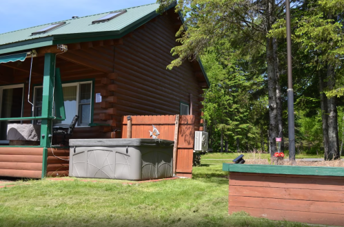 A log cabin with a green roof, hot tub, and garden area surrounded by trees on a sunny day.